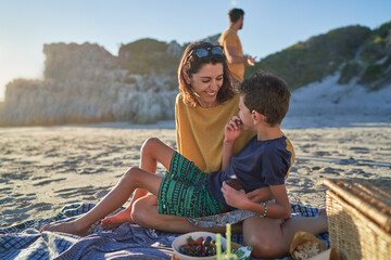 Happy mother and son eating on picnic blanket on sunny beach
