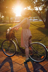 A woman with a bicycle, pink dress, and a hat watching the sunset in the park in summer.