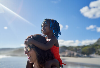 Happy mother carrying cute daughter on shoulders on sunny beach