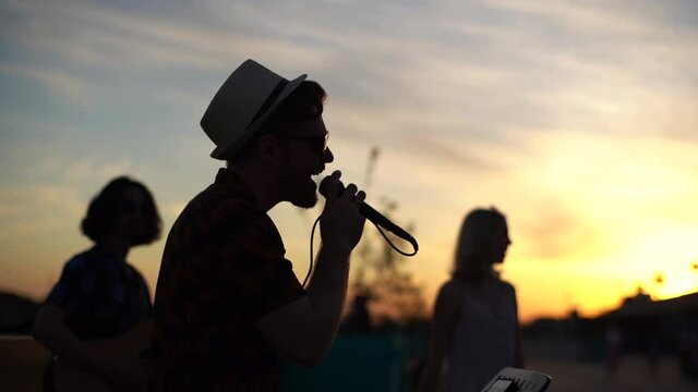 Silhouette of creative vocalist wearing hat and sunglasses singing in city embankment on background of warm sunlight during sunset. Talented young music group performing in street.