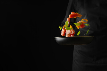 A beautiful composition of pieces of red fish, vegetables and herbs in a frying pan in a state of levitation. A cook in a black uniform holds a frying pan with fish, vegetables and herbs in his hand.