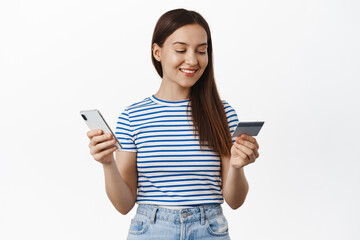 Online shopping. Young woman paying with credit card and mobile phone, smiling and looking relaxed, purchase smth in internet store, buying in smartphone application, standing over white background