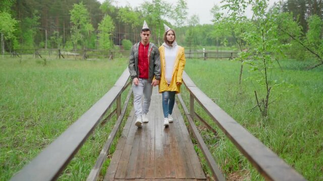 Sad unhappy young couple in raincoats and party hats walking down wooden path in countryside in rain in slow motion. Ruined dreams and broken expectations concept