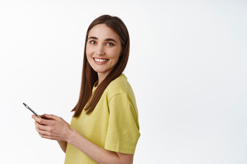 Naklejka premium Portrait of young candid girl, smiling natural, using smartphone, holding cellphone and looking happy at camera, chatting, standing against white background