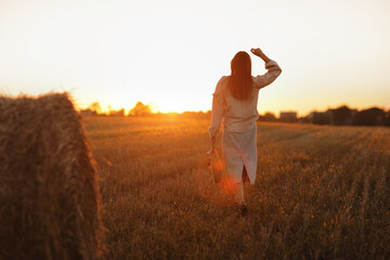young woman in the beautiful light of the summer sunset in a field is walking near the straw bales. beautiful romantic girl with long hair outdoors in field
