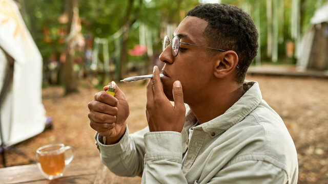 young afro man sitting in front of wooden table and lighting a joint