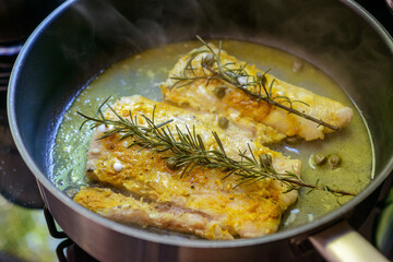 Fish fillet being fried in frying pan with rosemary and capers.