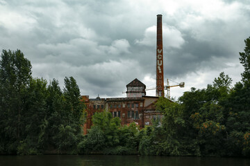 Fototapeta na wymiar an old German factory surrounded by green trees standing on a canal near the water. Cloudy weather and a little lyrical mood