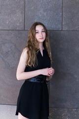 Medium vertical view of beautiful teenaged girl dressed in a black summer dress leaning against a stone background with demure expression