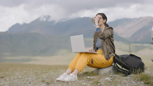 Freelancer works remotely while traveling on laptop while sitting near car in mountains. Young self-employed woman works in beautiful mountainous area