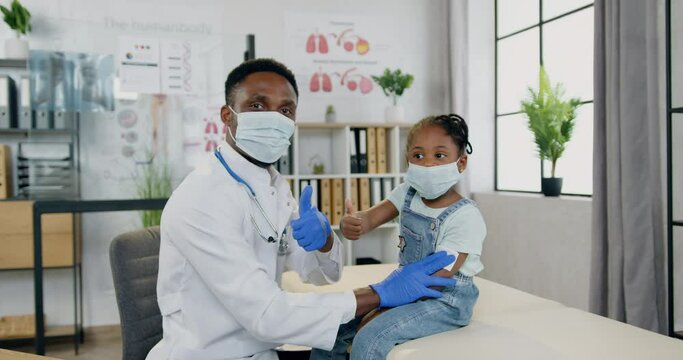 Handsome caring positive experienced black-skinned male doctor and cute small black-skinned girl both in protective masks posing on camera in modern medical office with thumbs up