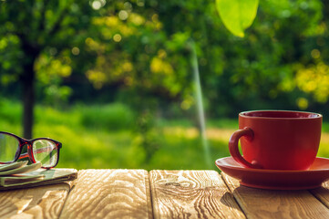 A cup of hot coffee and glasses with a newspaper on a wooden table in a green garden in the morning. Summer background