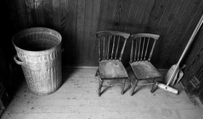 Somber looking scene of two old wooden children’s chairs sitting next to a garbage can and broom.
