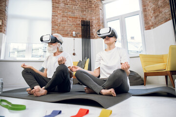 Relaxed aged man and woman wearing VR glasses while sitting in lotus position at bright living room. Active senior couple meditating at home using innovative headset.