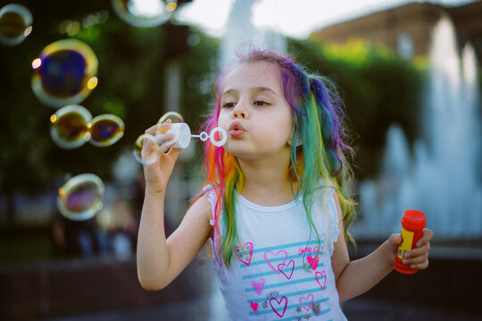 pretty little girl with colorful dyed hair blowing soap bubbles. Splashing city fountain is on background. Saint-Petersburg, Russia.Image with selective focus 
