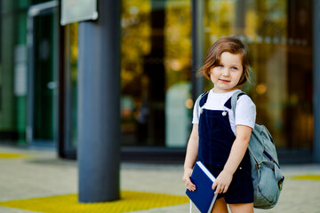 a beautiful Caucasian girl, a schoolgirl, is standing near the school, with a backpack and holding a textbook in her hands