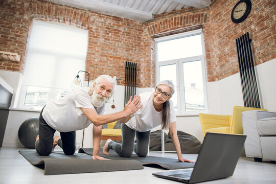 Happy senior man and woman in sport clothes giving high five while standing on yoga mat. Mature family watching tutorial on laptop for practicing yoga at home.