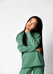 Thoughtful pensive asian preteen girl wearing green clothing, standing against studio background looking up and thinking on idea or problem making decision choosing solution touching chin by hand