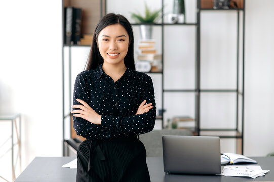 Portrait of successful confident pretty, young asian business woman or freelancer standing near work desk in office, wearing formal stylish clothes, arms crossed, looking at camera, smiling friendly