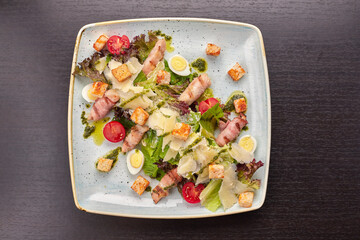Caesar salad with herbs chicken bacon eggs and tomatoes on a square light plate, on a dark background