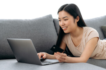 Online shopping. Positive beautiful young asian brunette woman lying on a couch, uses laptop and credit card to pay for online purchases and delivery, enters data, looks at the screen, smiles