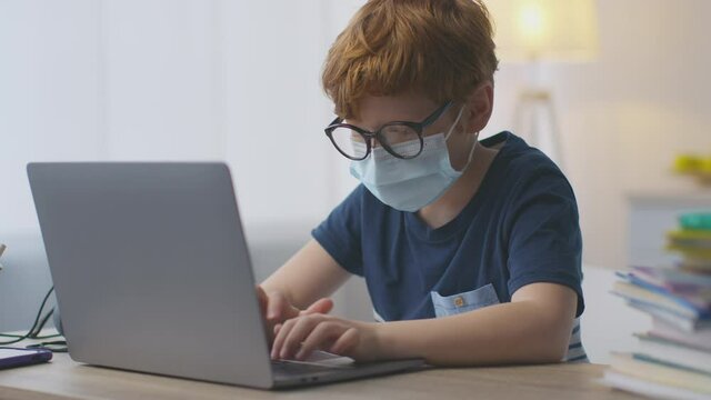 Quarantine measures and distant learning. Little redhead schoolboy wearing protective medical mack typing on laptop