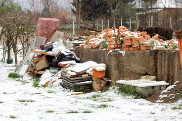 Bunch of various junk from construction material made of bricks and bathtub to broken windows and old sofa next to dilapidated family house concrete foundation surrounded with small trees and plants