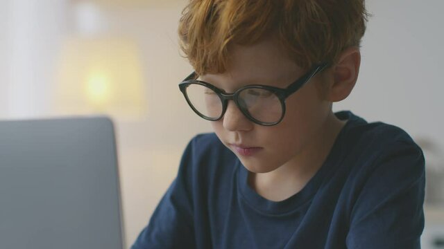 Kids development and upbringing. Llittle concentrated redhead schoolboy nerd studying at home on laptop, slow motion