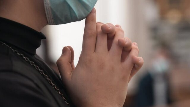 Nun in protective face mask crossing hands and praying in church during holy mass, close-up of religious sister's hands crossed on blurred church interior background. Christian in black cassock