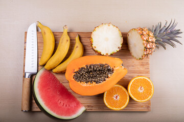 A cutting board with cut fruit on top and a knife, a papaya, a watermelon, a pineapple, an orange and bananas.