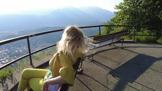 SLOW MOTION: Woman sitting on top of Lugano city by Lugano Lake in Switzerland. Swiss bench at aerial view lookout from Monte Bre Mount. Lugano cityscape with San Salvatore mount in Ticino canton.