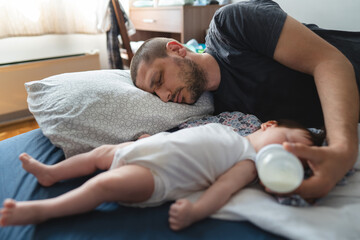 Exhausted man father lying by his three months old baby on the bed at home sleeping while holding...