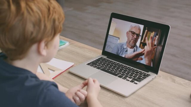 Online mathematics lesson. Little redhead schoolboy video chatting with male math teacher via laptop, tracking shot