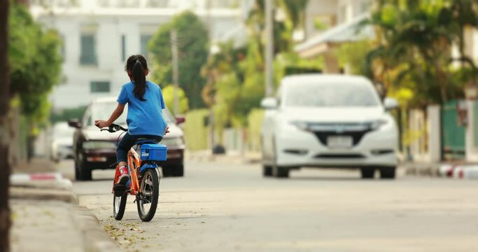 Backside of child girl riding a bicycle on street in the village with fun in slow motion shot. Kid leisure activity and sport concept.
