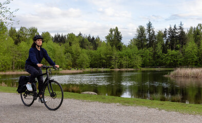 Adult Caucasian Woman Riding a Bicycle on a path by a lake in a modern city park. Spring Evening. Taken in Green Timbers Urban Forest, Surrey, Vancouver, British Columbia, Canada.