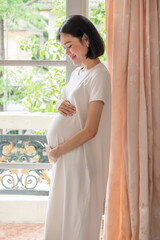 A pregnant Asian woman wearing casual clothes is standing smiling and looking her belly in a good mood.