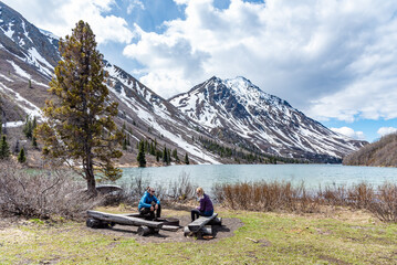 Fototapeta premium Hikers resting, sitting beside a isolated St Elias Lake in Kluane National Park during spring time, May with no other people. Snow capped mountains surrounding the scenic, Canadian view. 