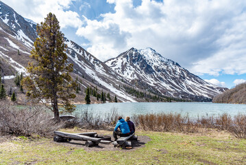 Woman and man couple sitting beside a lake in northern Canada with calm lake, blue sky and snow capped mountains. Scenic, iconic Canadian travel, tourism view. 