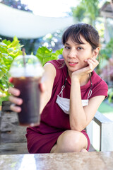 Fototapeta premium Asian women have masks hanging around their necks. Sit back and relax holding a glass of Black Coffee iced, delicious, refreshing, thirsty. look forward to a hot day In the garden of a coffee shop.