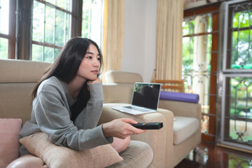 Young adult asian woman using remote tv for watching online movie at home in living room.