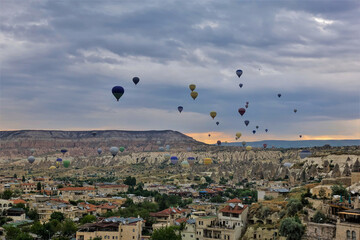 Flying over Cappadocia at dawn. In the cloudy sky, over the village, colorful balloons. The horizon is illuminated in orange. In the distance is a picturesque mountain with a flat top. Turkey