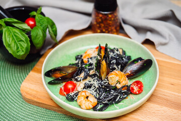 Black fettuccine pasta with shrimp, oysters, parmesan cheese, cherry tomatoes in a white wine sauce on a green plate and a wooden board on a background of gray napkin, paprika and basil leaves