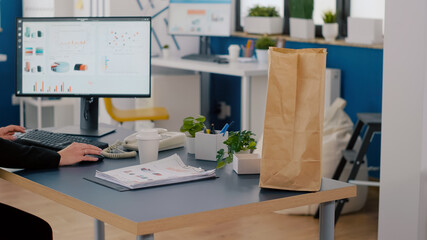 Close-up of entrepreneur woman holding delivery food order package putting meal bag on table during lunch break in company office. Executive manager typing at marketing statistics on computer