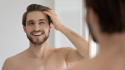 Happy handsome young guy combing smooth brown hair with fingers at mirror in bathroom, looking at...