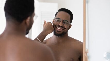 Fototapeta na wymiar Happy handsome mixed race Black guy in glasses touching soft stubble, satisfied stylish trimmed beard, barber work, aftershave balm, lotion. Metrosexual young man looking in mirror with toothy smile