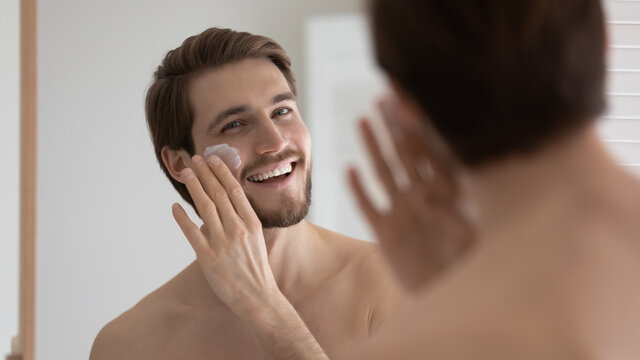 Happy handsome metrosexual guy applying sunscreen on face at mirror, spreading moisturizing collagen cream on under eye skin, reducing bags, wrinkles, protecting from sun. Male skincare concept - Powered by Adobe