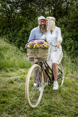 Obraz na płótnie Canvas blonde woman with white dress and her boyfriend or husband posing with bicycle with beautifully decorated flower basket in nature and are happy and in love, nature concept, flowers, delivery service