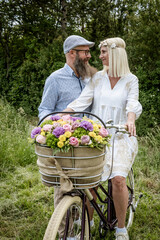 blonde woman with white dress and her boyfriend or husband posing with bicycle with beautifully decorated flower basket in nature and are happy and in love, nature concept, flowers, delivery service