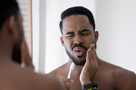 Stressed upset mixed raced Black man feeling pain and discomfort while brushing teeth in bathroom, holding toothbrush, touching cheek with painful grimace. Toothache, dental problem concept