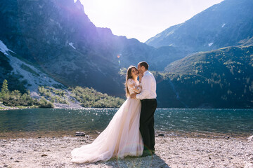 An elegant couple on a sunny day near a mountain lake, kissing, walking after the ceremony